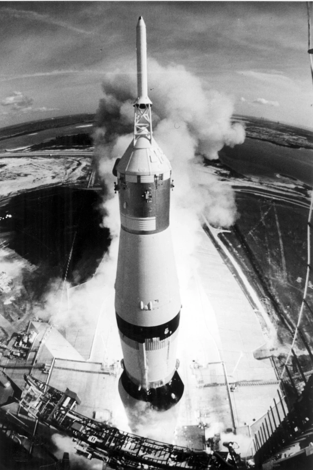 Saturn V rises beside its launch tower above a bright exhaust plume.